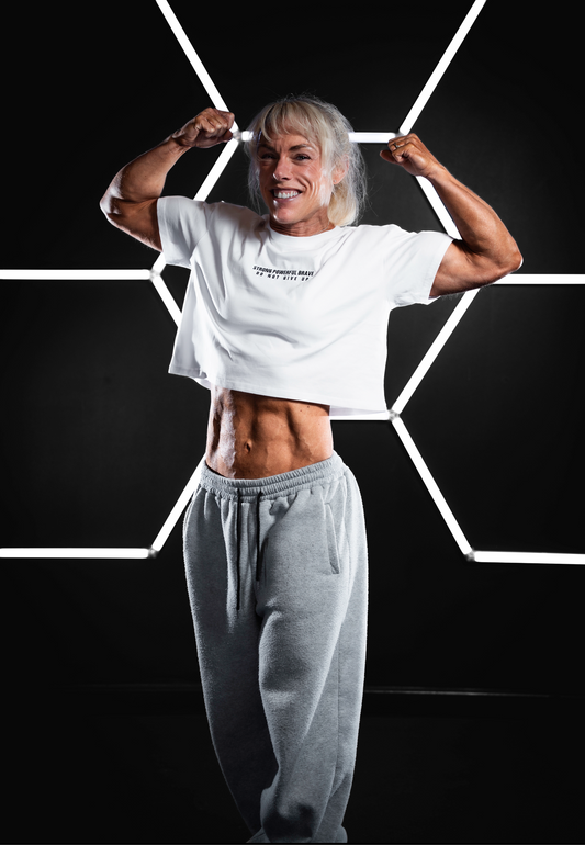 Female bodybuilder wearing a white SPB oversized cropped T-shirt with the “STRONG POWERFUL BRAVE – DO NOT GIVE UP” slogan printed on the chest. She stands confidently in front of a hexagonal light installation, flexing both arms to show muscular definition. Paired with grey sweatpants. The setting is bold and futuristic, emphasizing strength and empowerment.
