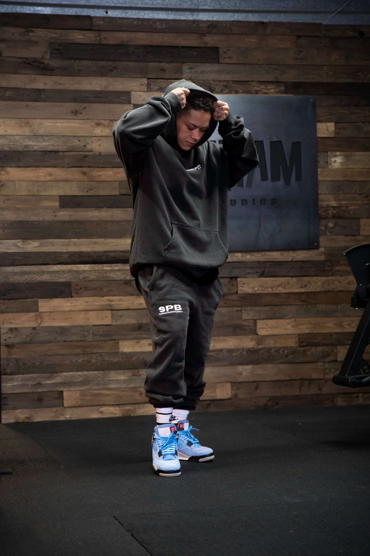 SPB branded oversizes hoodie and bottoms, standing infront of a wooden wall.