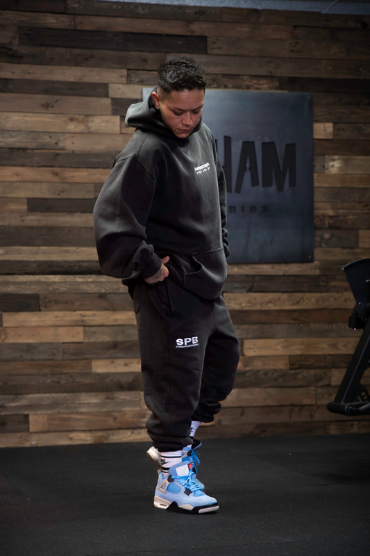 A person wearing black oversized SPB joggers, a hoodie, and blue and white sneakers, standing in front of a wooden backdrop.