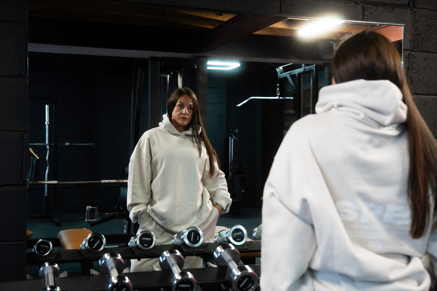 Female model wearing a 2XL SPB oversized hoodie and XL bottoms in Faded Bone, facing away in a raw gym setting. The shot highlights the relaxed fit and powerful presence of SPB’s bold, inclusive streetwear.