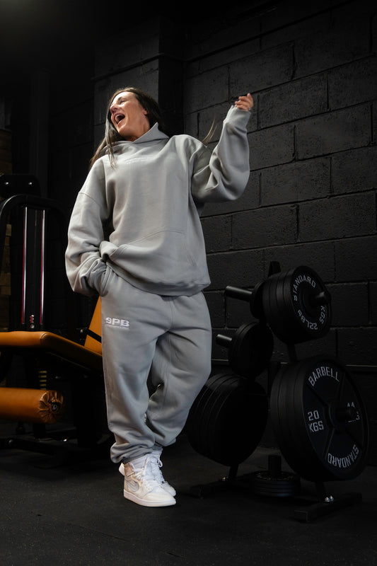 Person wearing a gray SPB oversized hoodie with kangaroo pocket and matching bottoms, standing in a gym with exercise equipment in the background.