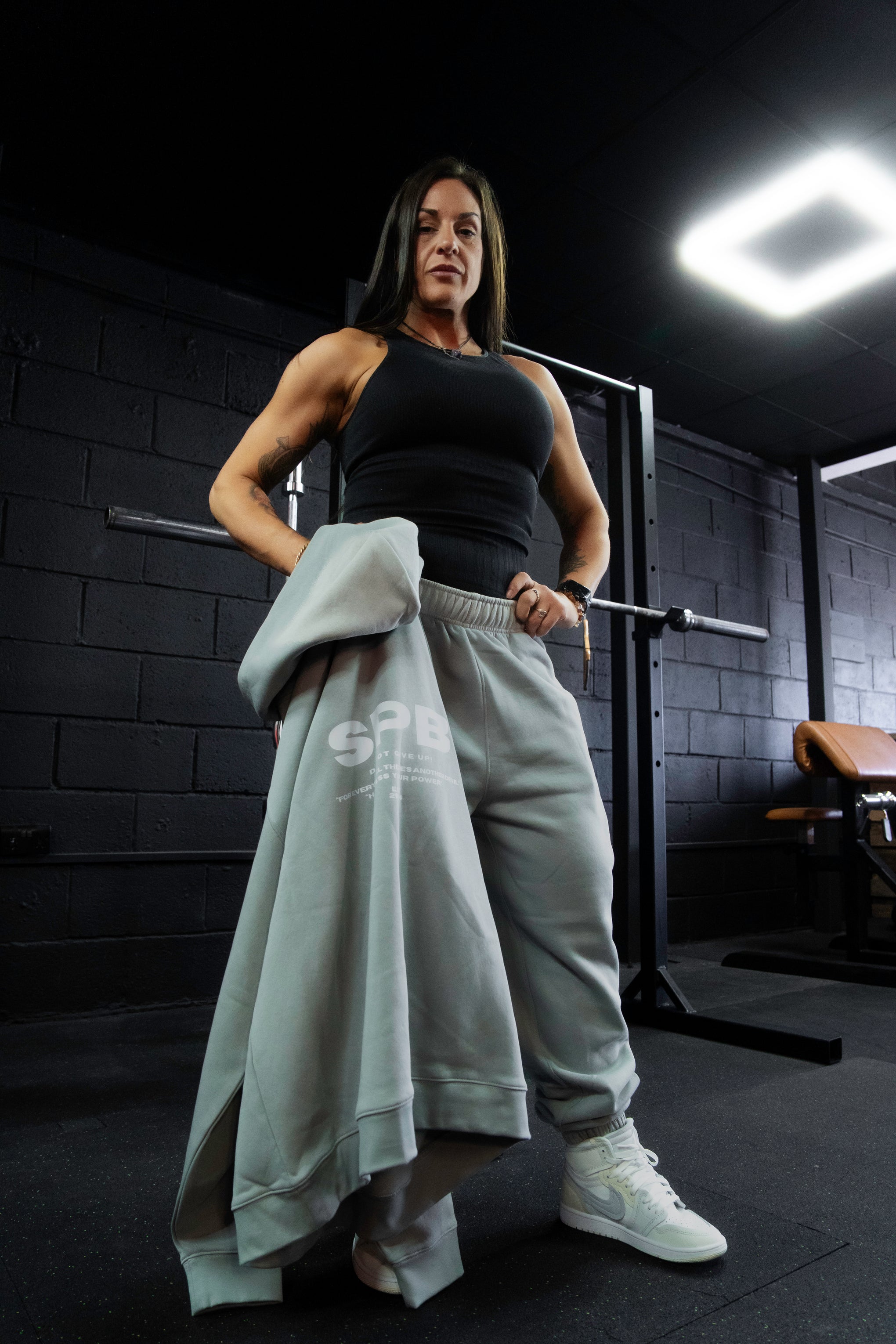 Person wearing gray SPB bottoms, black top and holding gray SPB hoodie in a gym setting.