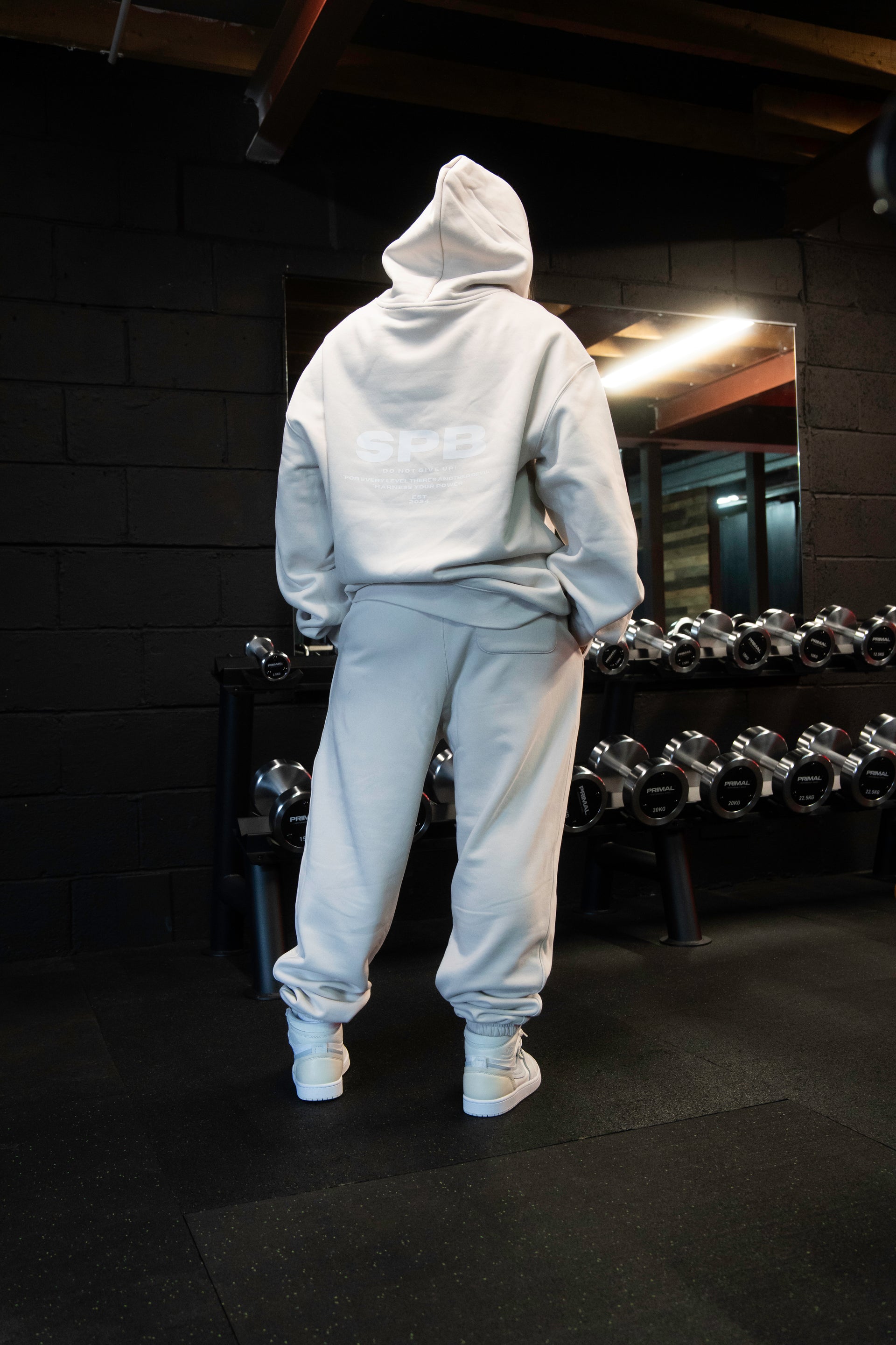 Female model wearing a 2XL SPB oversized hoodie and XL bottoms in Faded Bone, facing away in a raw gym setting. The shot highlights the relaxed fit and powerful presence of SPB’s bold, inclusive streetwear.
