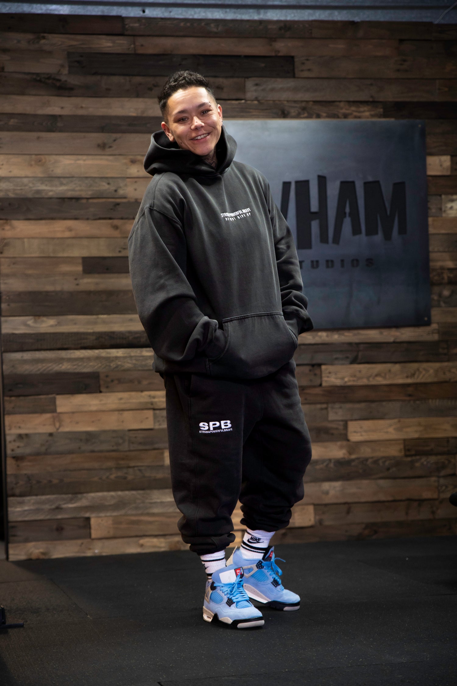 A person wearing a black oversized SPB hoodie and bottoms, standing in front of a wooden panel wall.