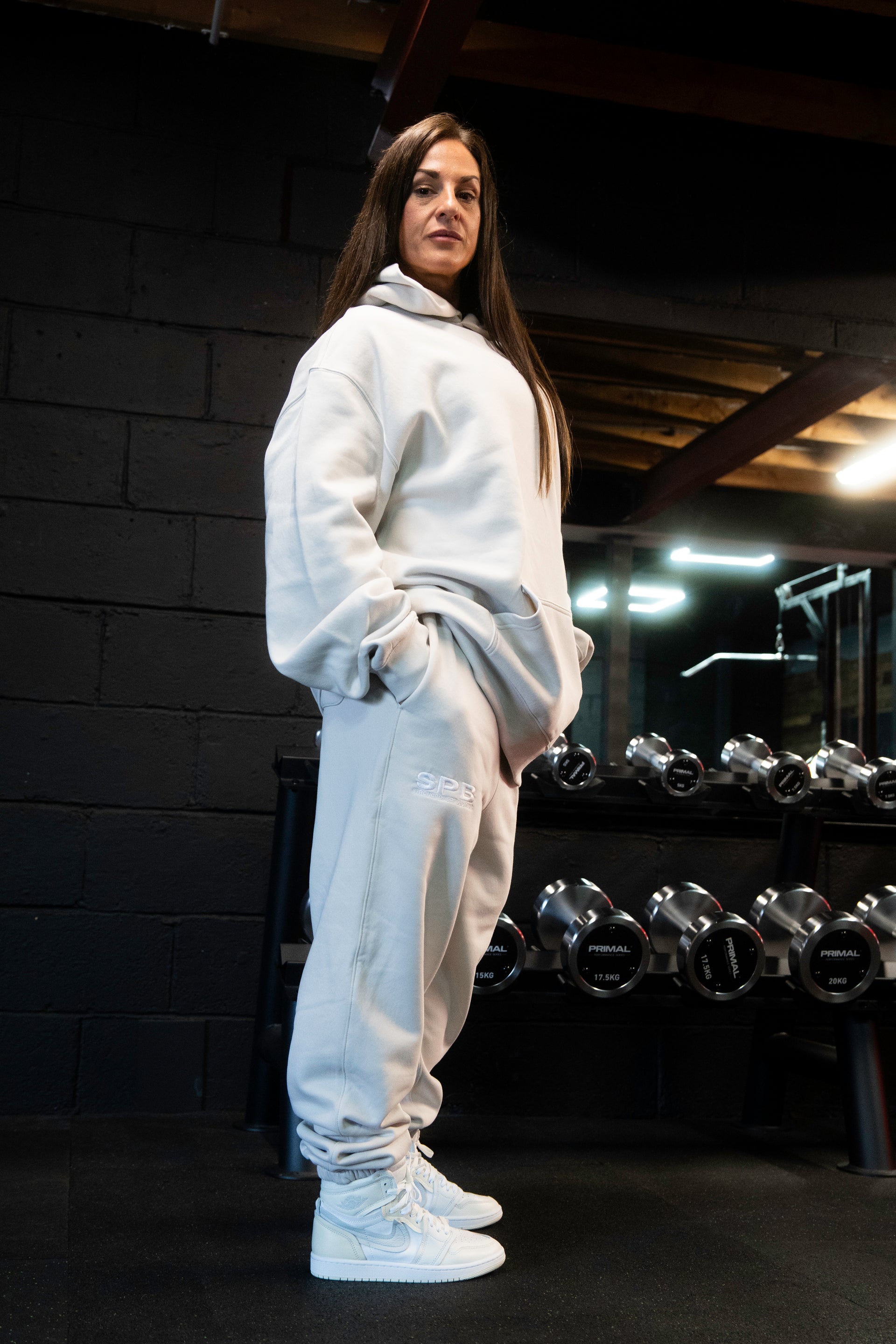 Female model wearing a 2XL SPB oversized hoodie and XL bottoms in Faded Bone, facing to the right in a raw gym setting. The shot highlights the relaxed fit and powerful presence of SPB’s bold, inclusive streetwear.
