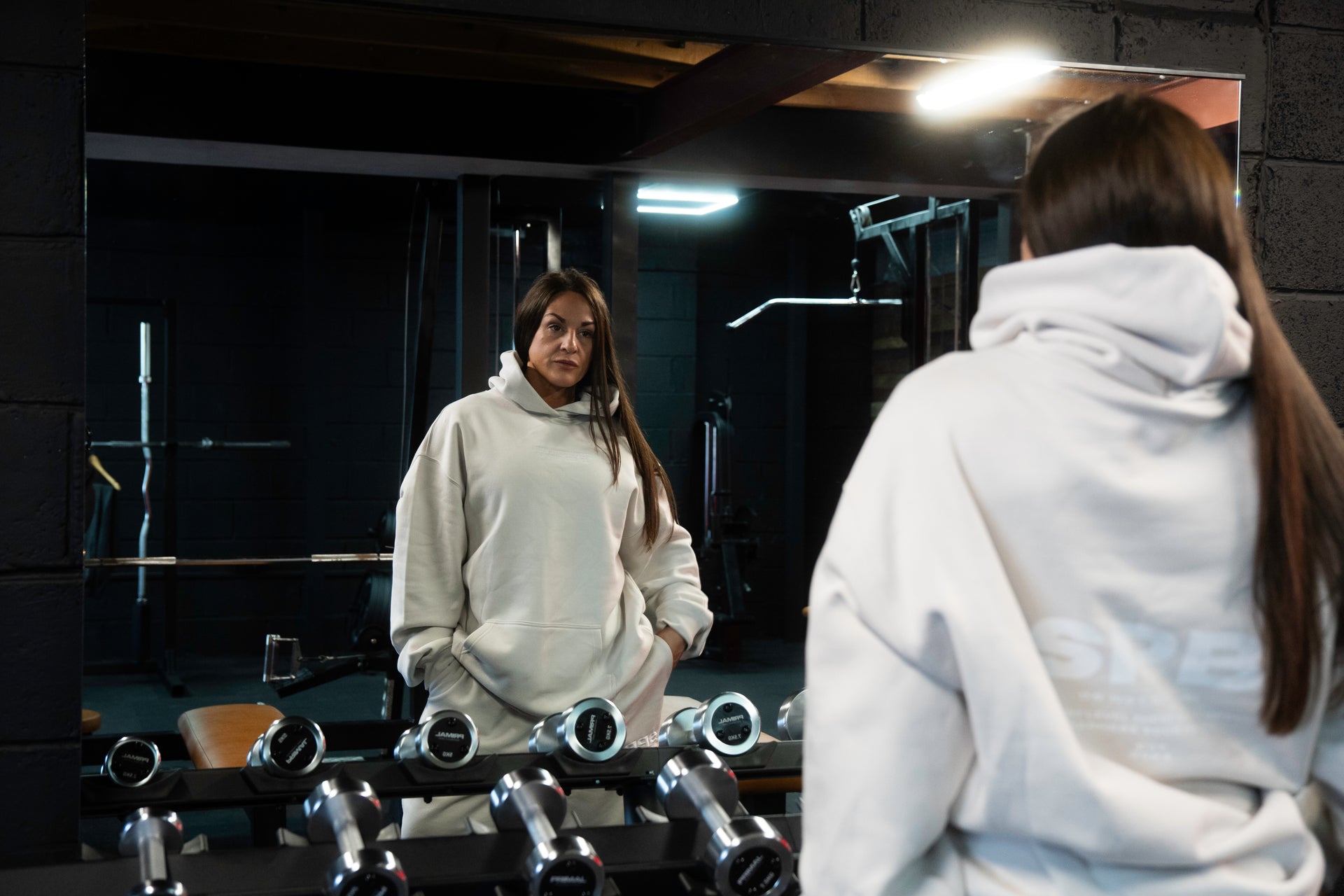 Female model wearing a 2XL SPB oversized hoodie and XL bottoms in Faded Bone, facing away in a raw gym setting. The shot highlights the relaxed fit and powerful presence of SPB’s bold, inclusive streetwear.
