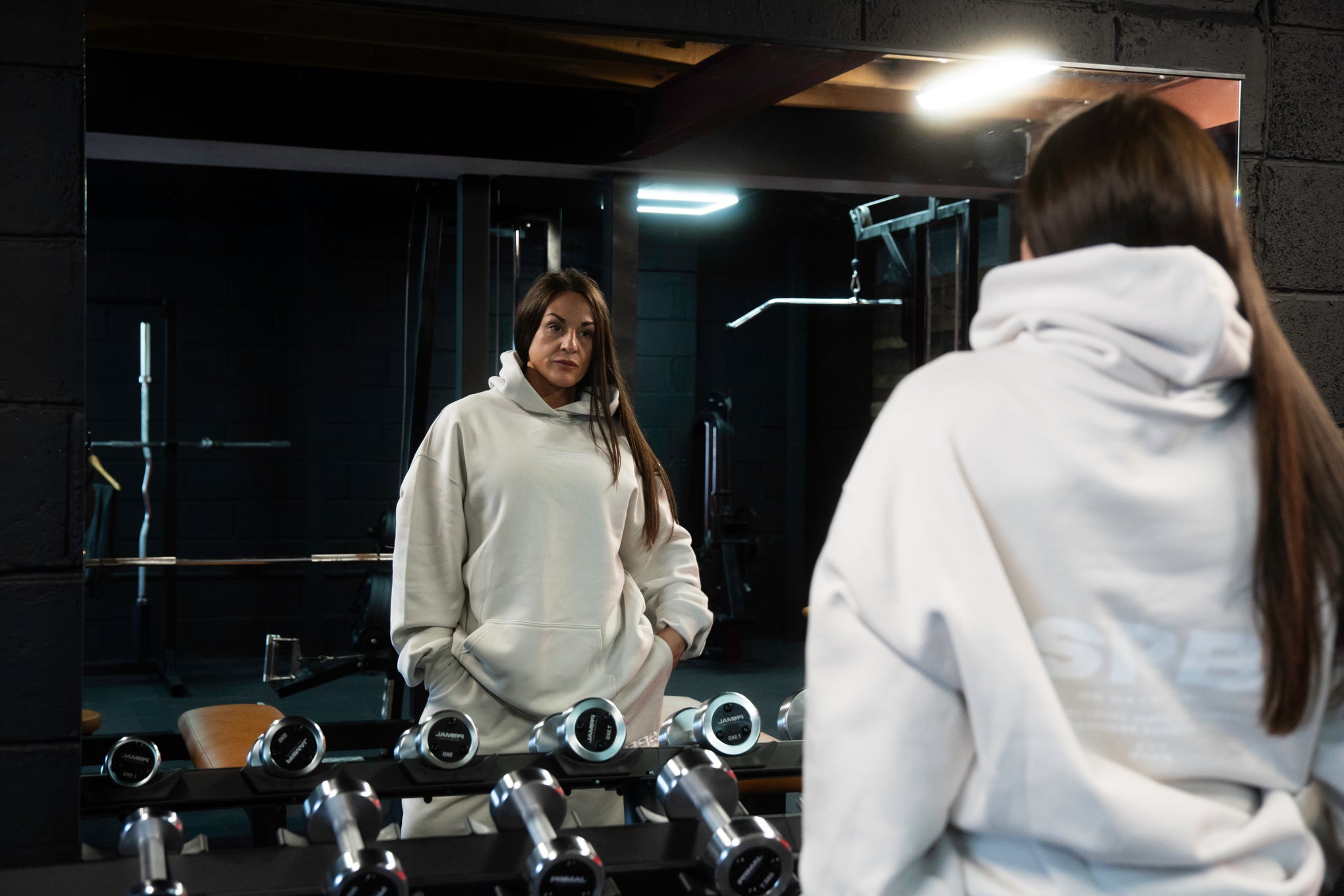 Female model wearing a 2XL SPB oversized hoodie and XL bottoms in Faded Bone, facing away in a raw gym setting. The shot highlights the relaxed fit and powerful presence of SPB’s bold, inclusive streetwear.
