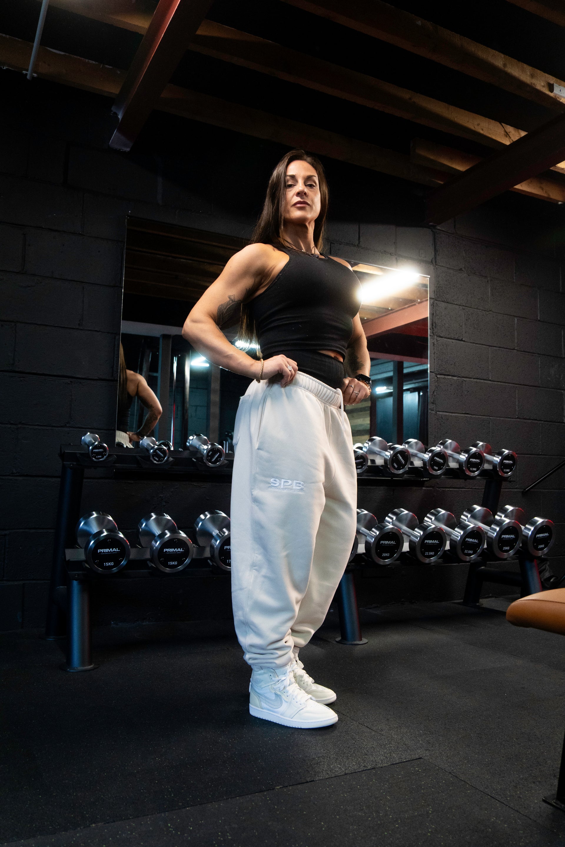 A person wearing gray SPB oversized joggers, a black top, and blue and white sneakers, standing in front of dumbells.