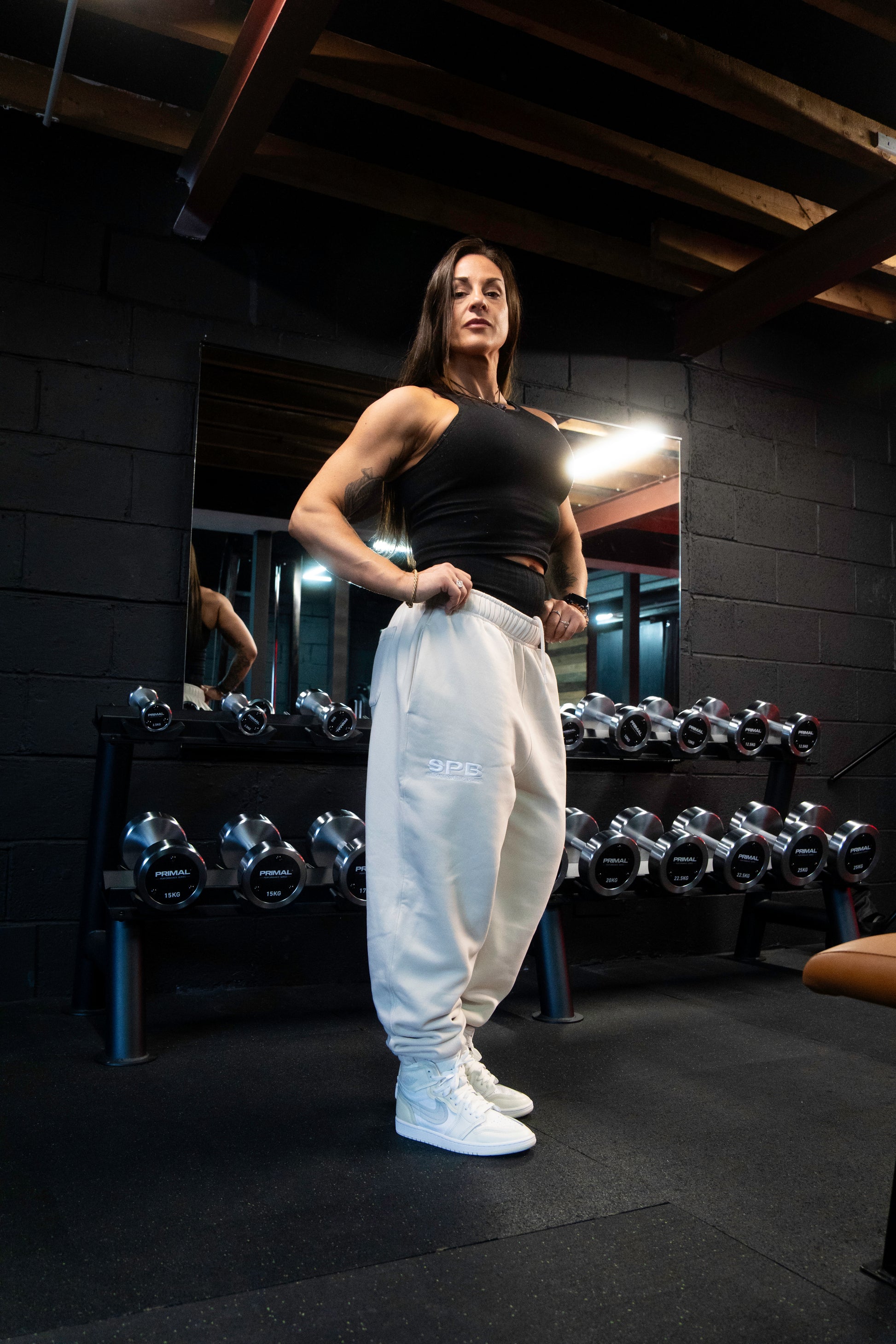A person wearing gray SPB oversized joggers, a black top, and blue and white sneakers, standing in front of dumbells.