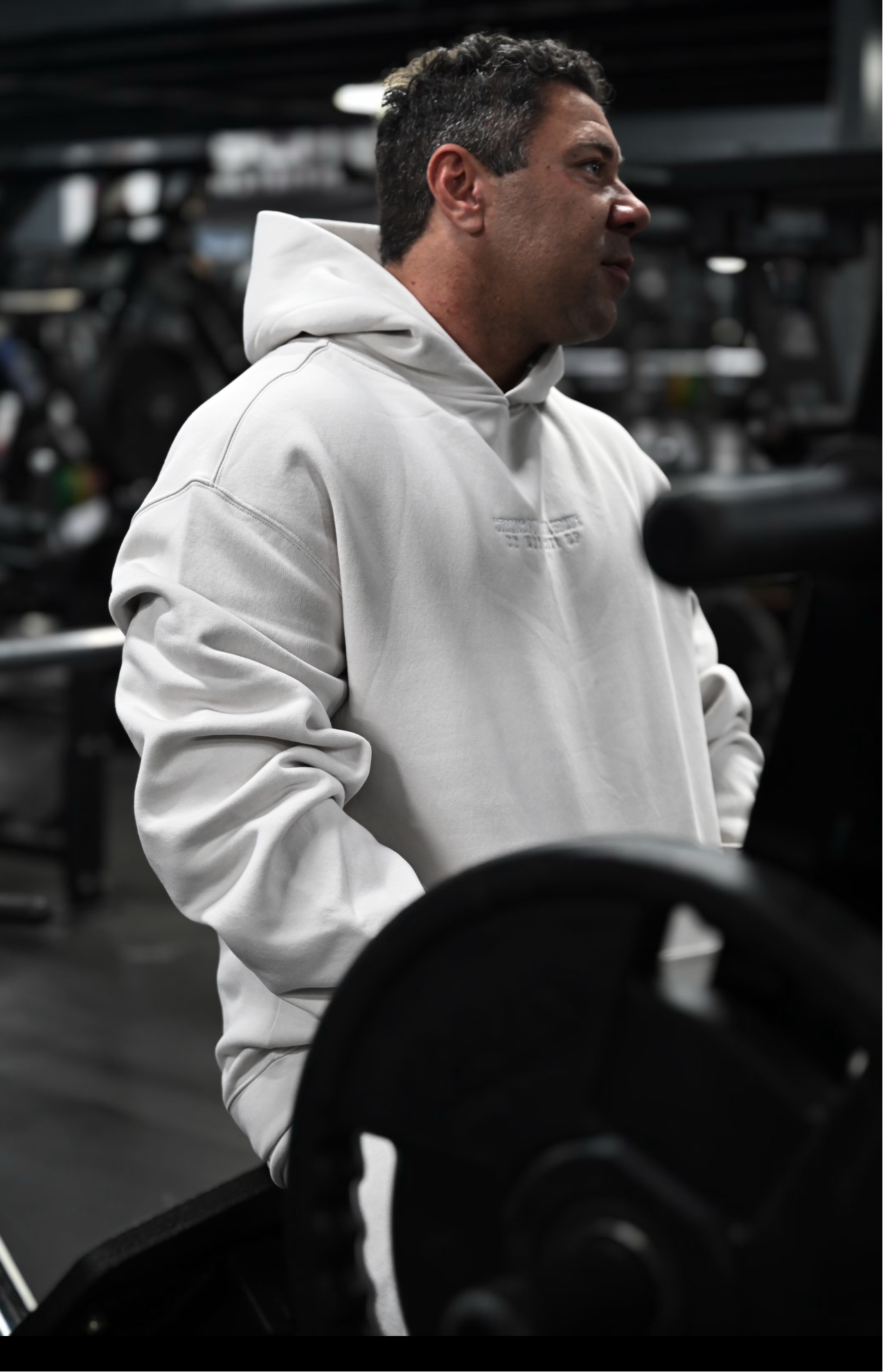Male model wearing a 2XL SPB oversized hoodie and XL bottoms in Faded Bone, sitting confidently in the foreground, looking off to the side. Urban, unfiltered setting showcasing SPB’s bold, inclusive streetwear.
