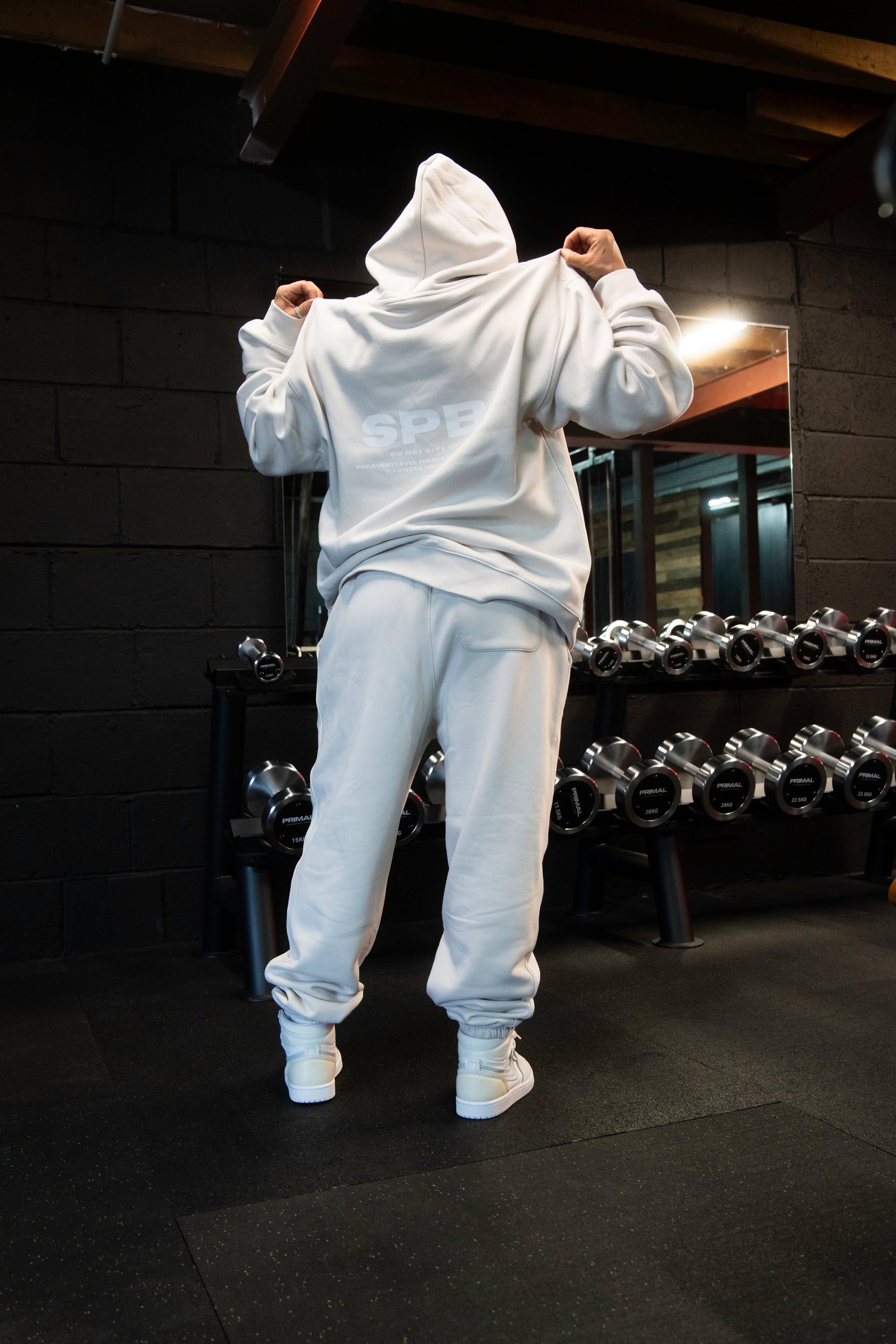 Female model wearing a 2XL SPB oversized hoodie and XL bottoms in Faded Bone, facing away in a raw gym setting. The shot highlights the relaxed fit and powerful presence of SPB’s bold, inclusive streetwear.
