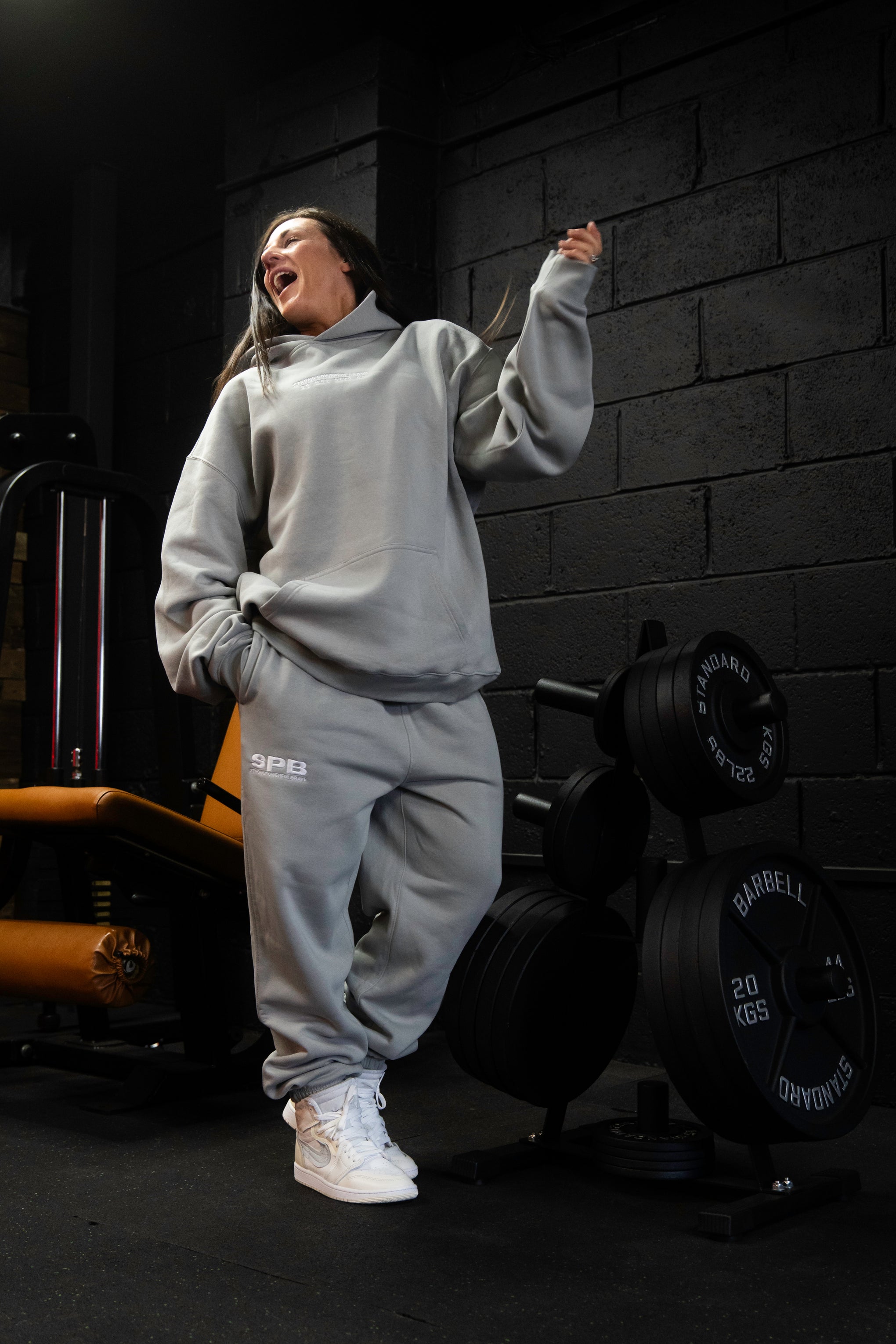 Person wearing a gray SPB oversized hoodie with kangaroo pocket and matching bottoms, standing in a gym with exercise equipment in the background.