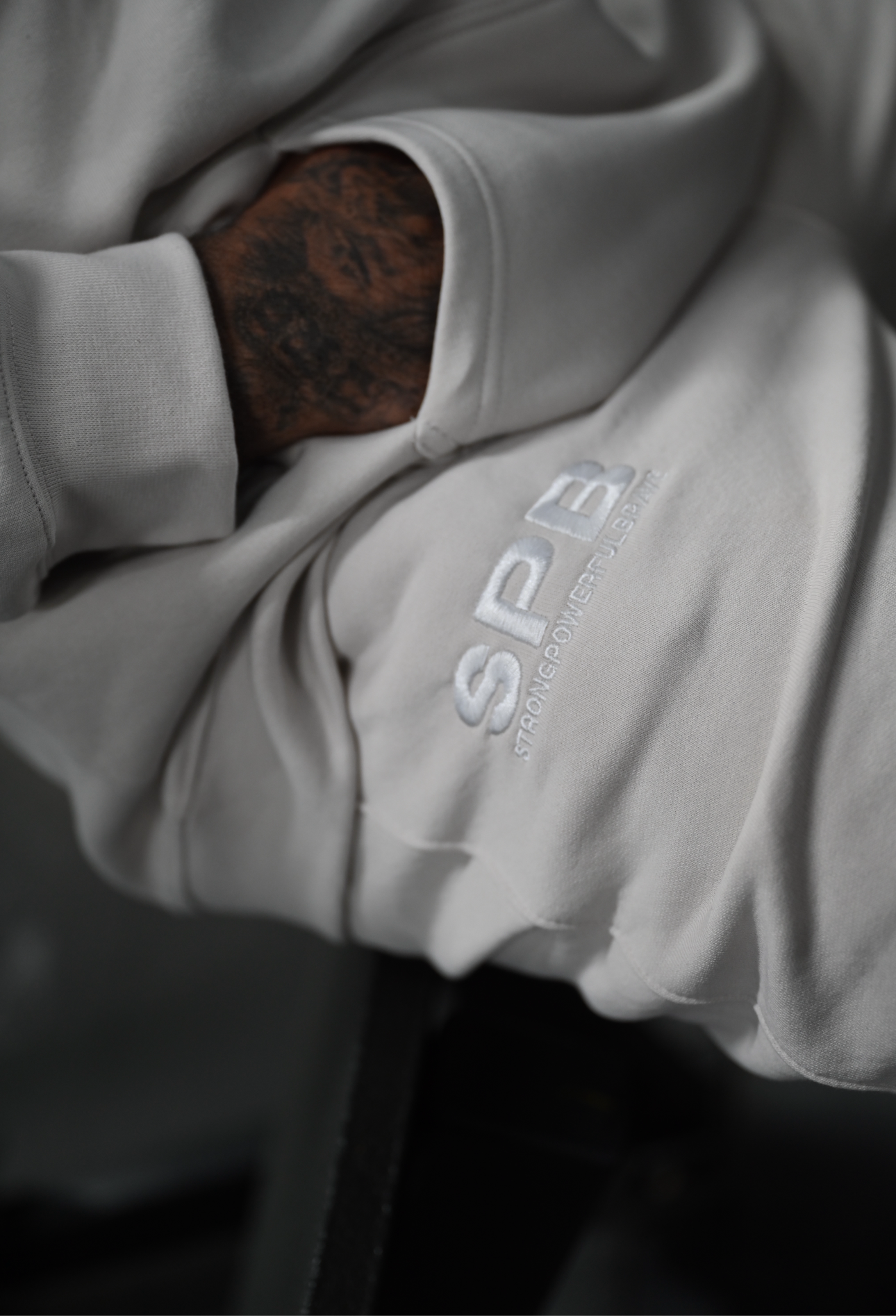 Male model wearing a 2XL SPB oversized hoodie and XL bottoms in Faded Bone, sitting confidently in the foreground. A close up of the SPB logo. 