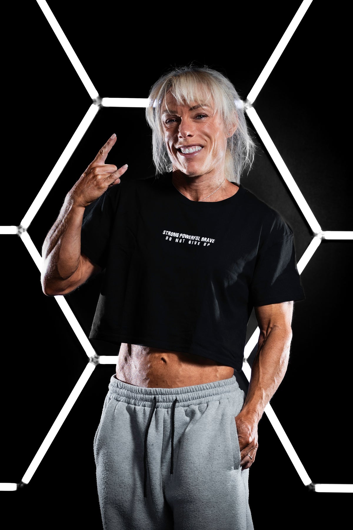 Female bodybuilder wearing a black SPB oversized cropped T-shirt with the “STRONG POWERFUL BRAVE – DO NOT GIVE UP” slogan on the chest. She smiles and throws a rock hand gesture, standing in front of the same geometric light background. Paired with grey sweatpants. The image radiates confidence, resilience, and a rebellious energy.