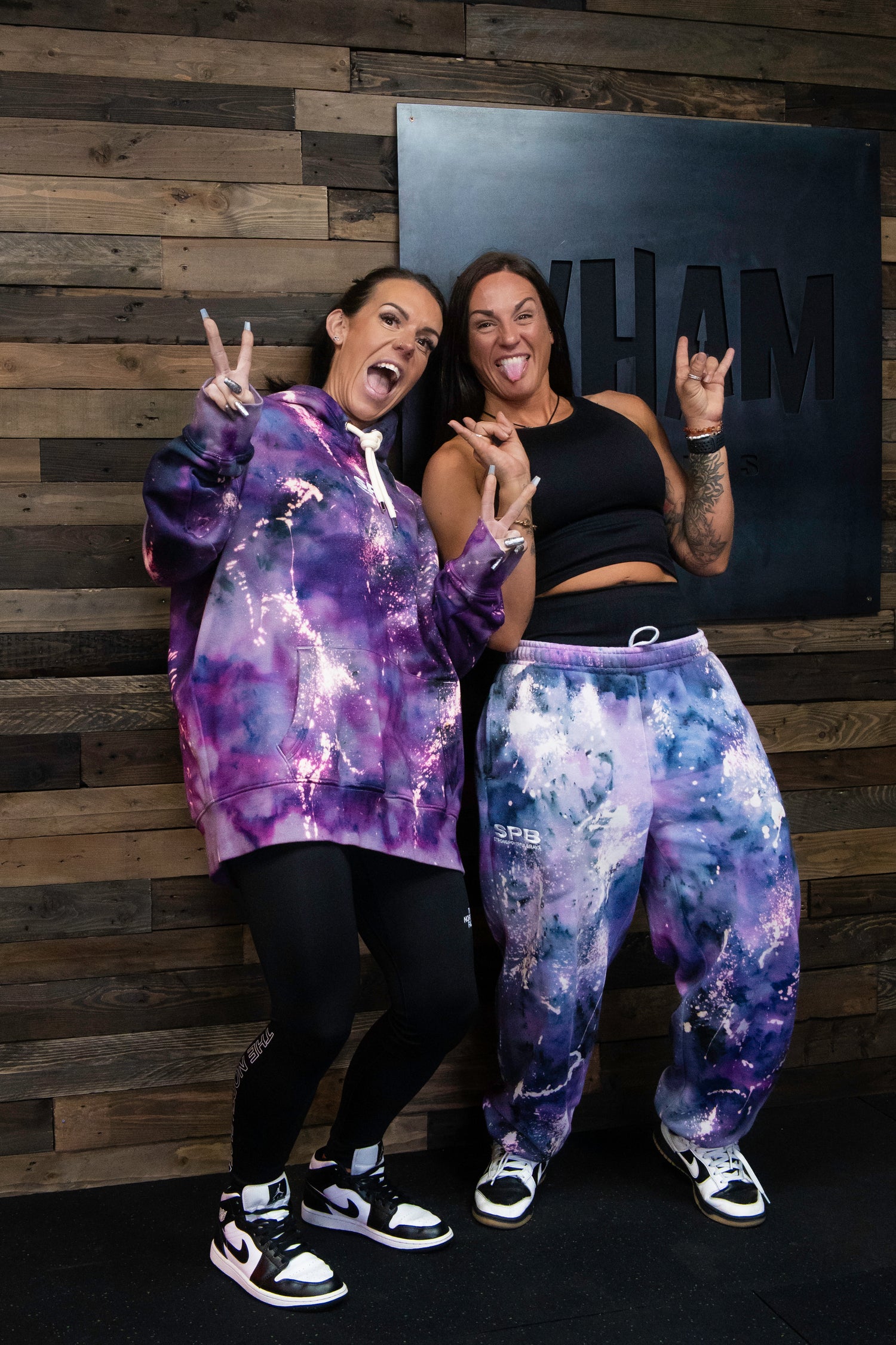 Two people wearing purple tie-dye SPB clothing, one is wearing a hoodie and the other bottoms in front of a wooden wall.